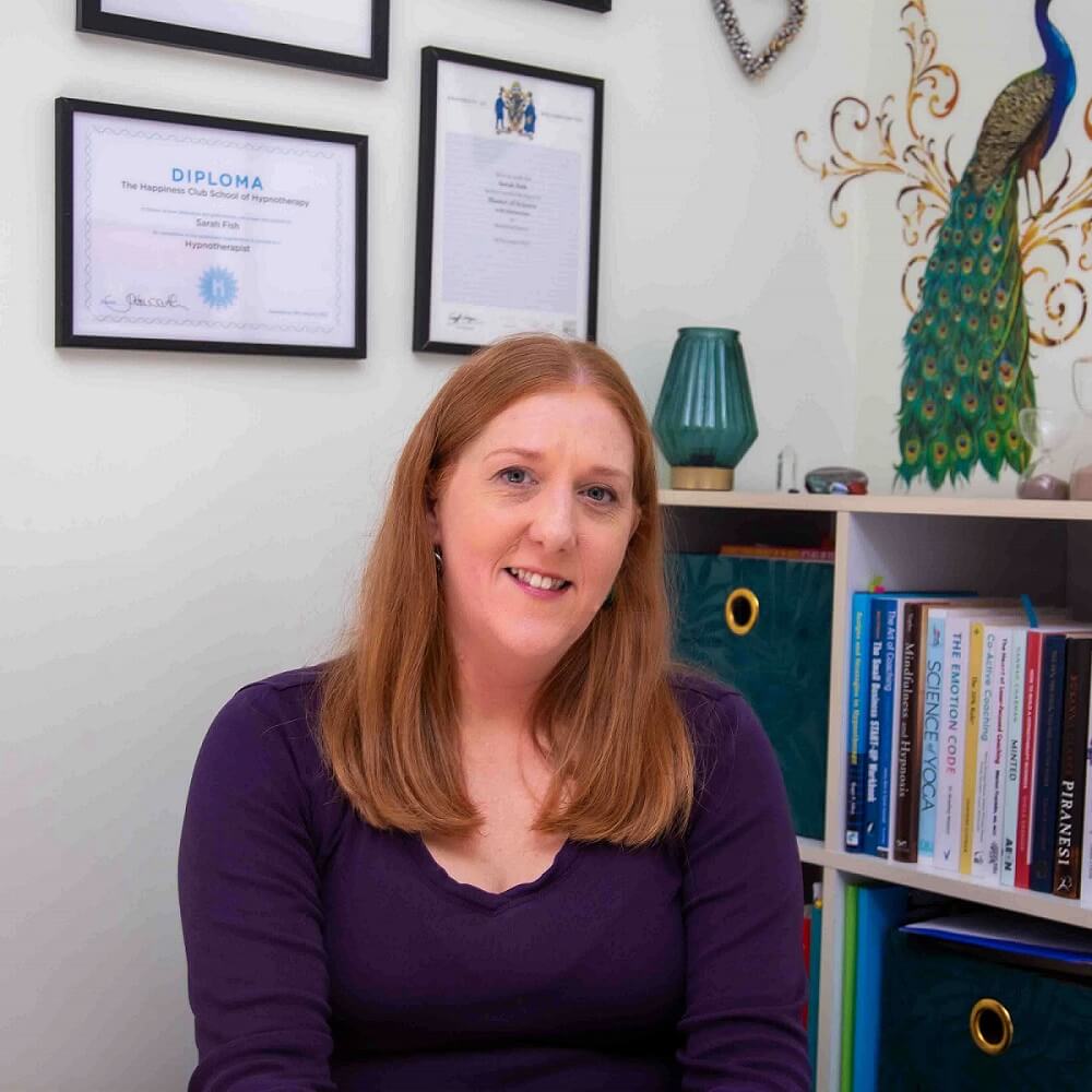 Sarah wears a dark purple top and has her ginger hair down. She sits facing the camera with a smile, in front of her book case and framed certificates that are on the wall behind her. On the wall there is also an image of a peacock in green and blue.